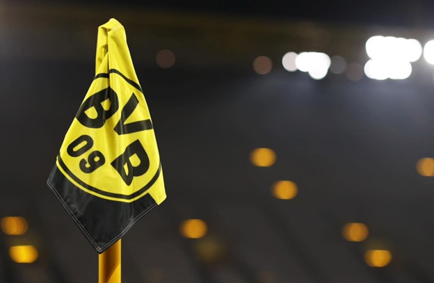 A bright yellow corner flag featuring the black logo ‘BVB 09’ waves in the foreground, with a blurred stadium and lights in the background.